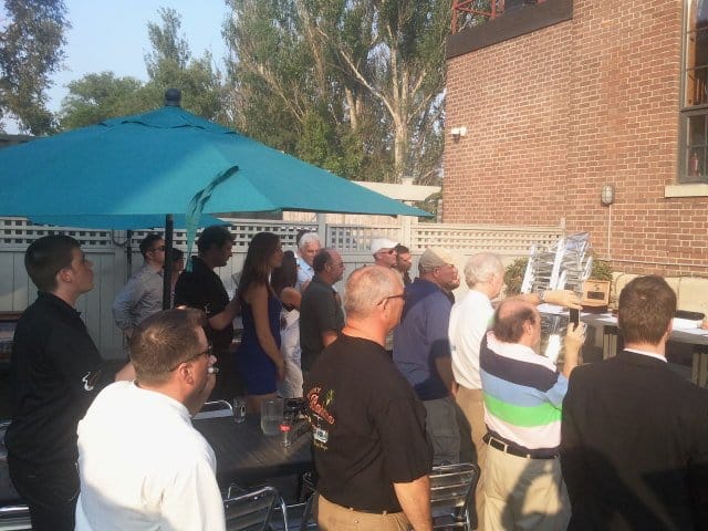 group of people standing listening to someone talk, brown brick wall, teal umbrella, green trees in the background